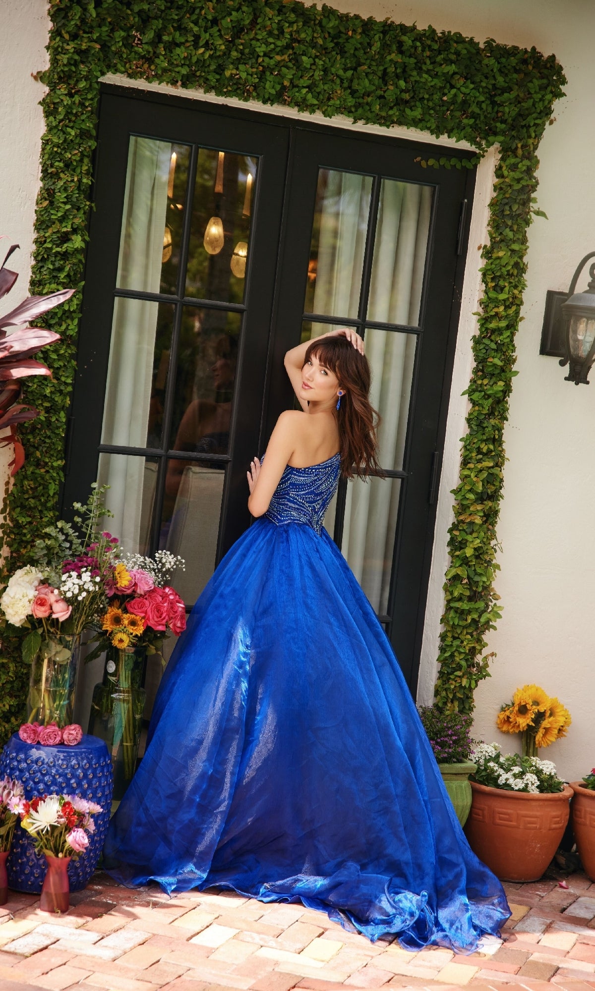 A woman in the Ava Presley 29258 one-shoulder prom ball gown stands outside by glass doors, surrounded by pots of colorful flowers and lush greenery.